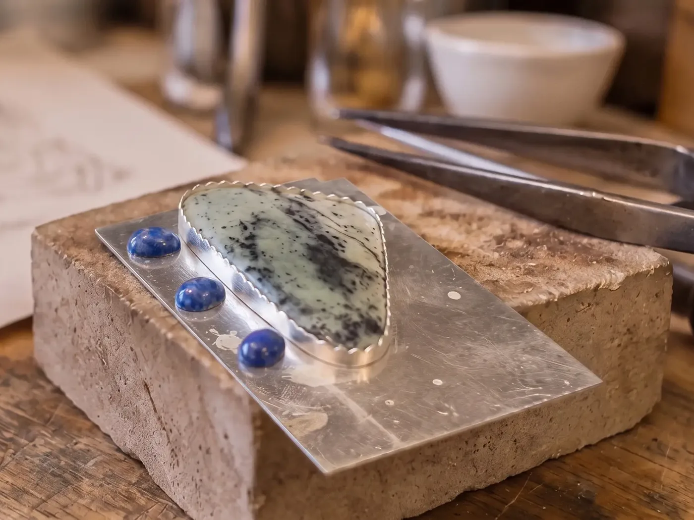 A green stone in a hand-cut bezel beside three lapis cabochons, on a soldering block at the bench