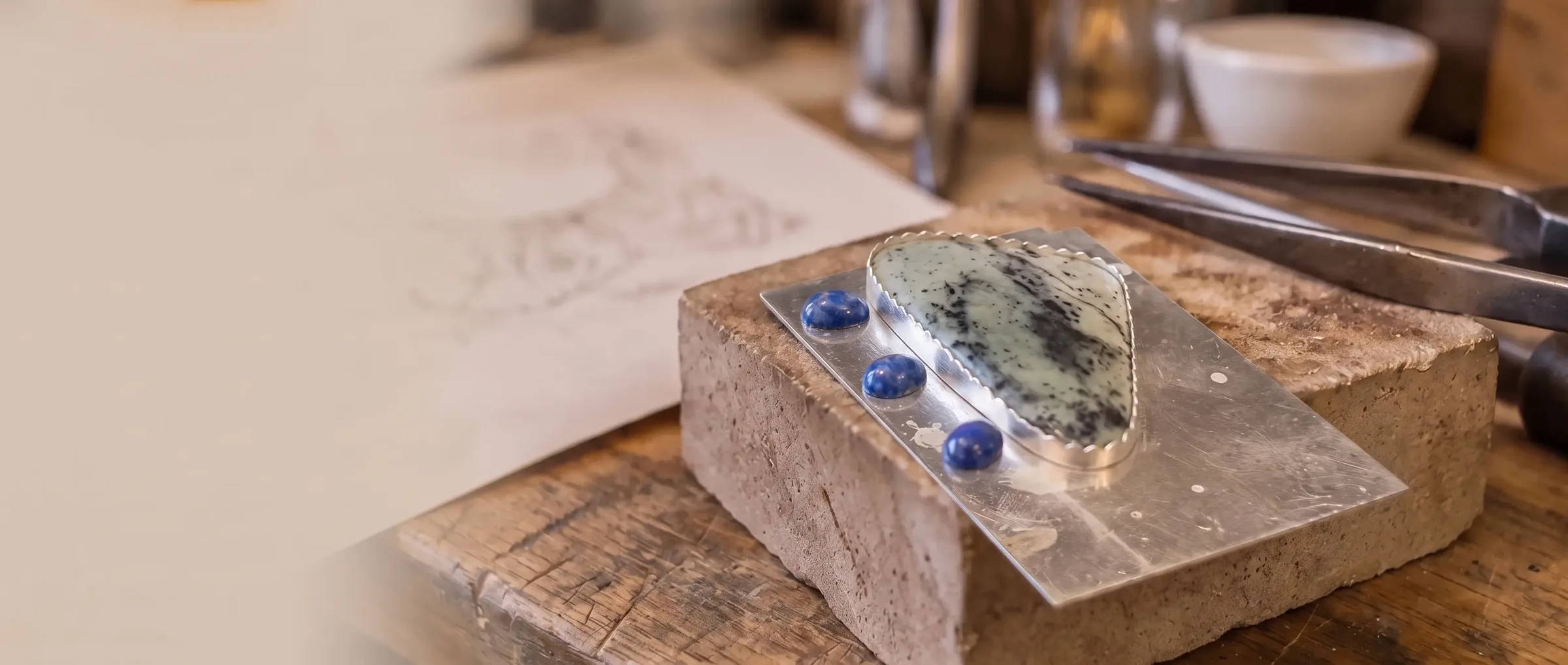 A green stone in a hand-cut bezel beside three lapis cabochons, on a soldering block at the bench
